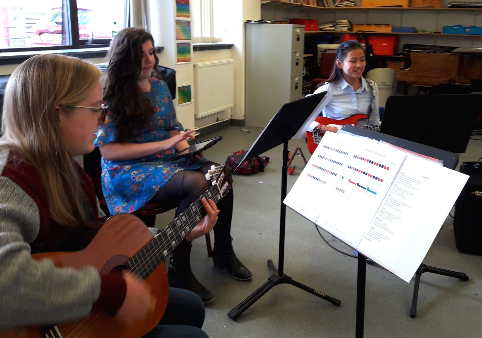 3 women play in a band using Figurenotes notation. 1 on guitar, 1 on iPad, and 1 on bass guitar