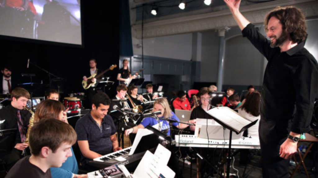 A smiling conductor raises his hand to direct a stage full of young musicians