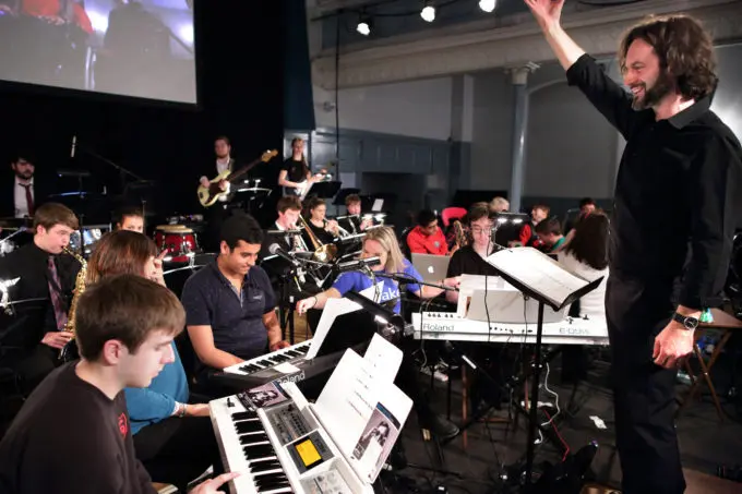 A group of young musicians on stage with a male conductor, arm raised.