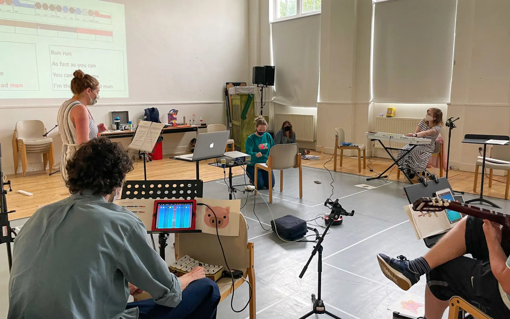 A group of musicians in a hall. A narrator is standing; seated woman on chimes; seated woman on keys; seated man with a pig flashcard, chimes, and an ipad showing Thumbjam. The edge of a guitar and its player can also be seen.