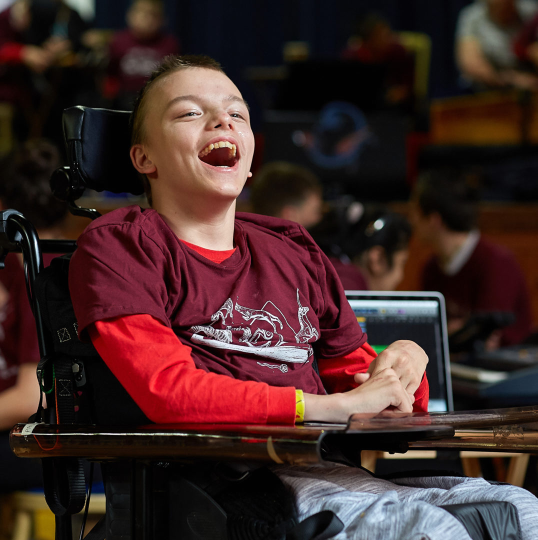 Smiling boy with hands together on a table. He is sitting in a wheelchair wearing a maroon tshirt.