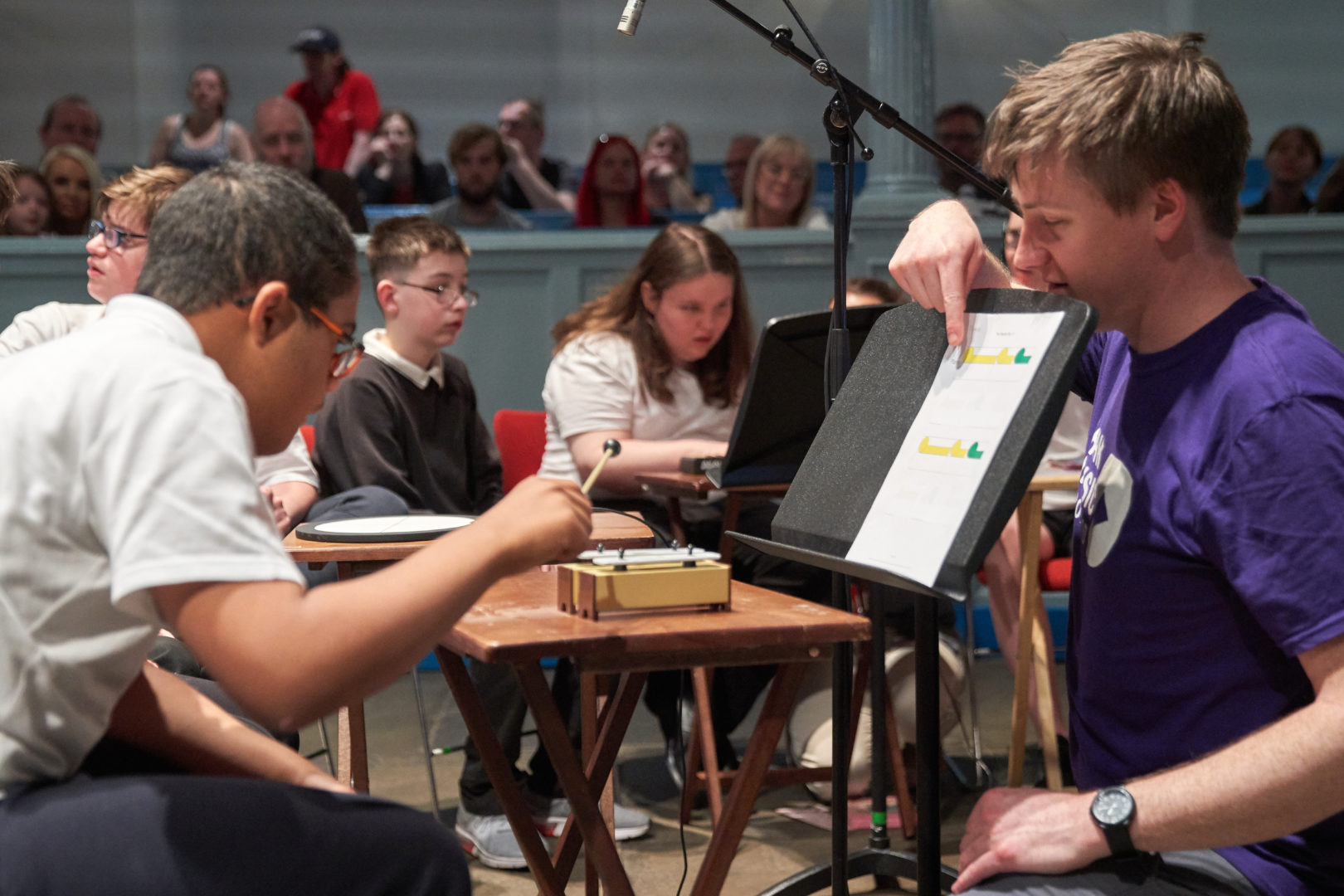 Young person at music concert playing chime bars with Figurenotes stickers. Tutor supporting young person by pointing to Figurenotes score.