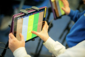 Two tablets are being played as a musical instrument on a stand with Figurenotes colours on the screen. Young persons hands are visibly playing the tablets. 