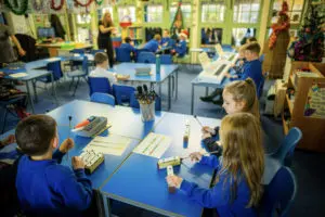 Classroom scene from a corner with students playing chime bars at tables with scores of colourful notation in front of them and a teacher in the middle of the classroom directing.