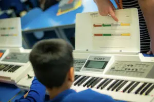 The back of a school child's head, brunette hair, playing a keyboard with colourful notation on a score in front of them. Teachers hand visible aiding the young person by pointing at the colourful score.