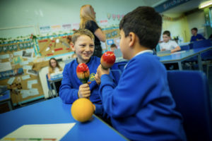 Two school children, one blonde, one with dark hair, facing each other playing wooden maracas and smiling at a school desk.