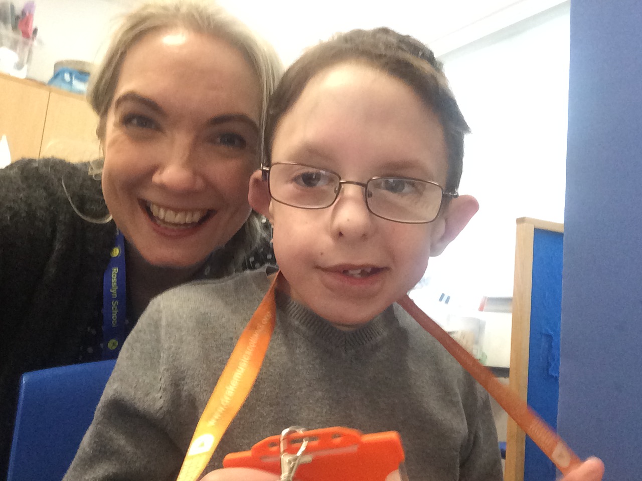 A smiling blonde woman with her head close to a smiling little boy in glasses. Their faces are side by side.