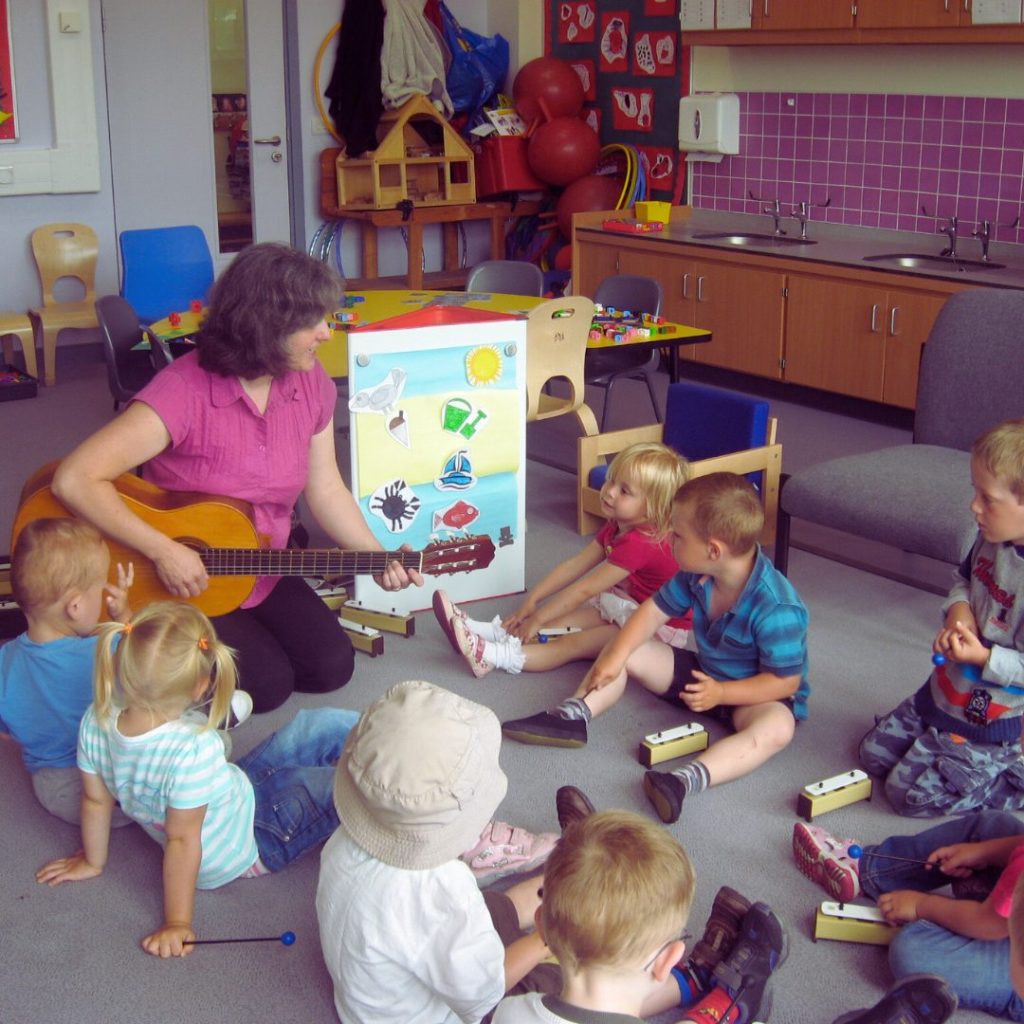 A woman plays guitar to a group of small children. They each have chime bars and are sitting on the floor. There is a board with a seaside image on it.