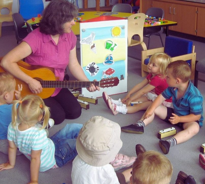 A woman plays guitar to a group of small children. They each have chime bars and are sitting on the floor. There is a board with a seaside image on it.