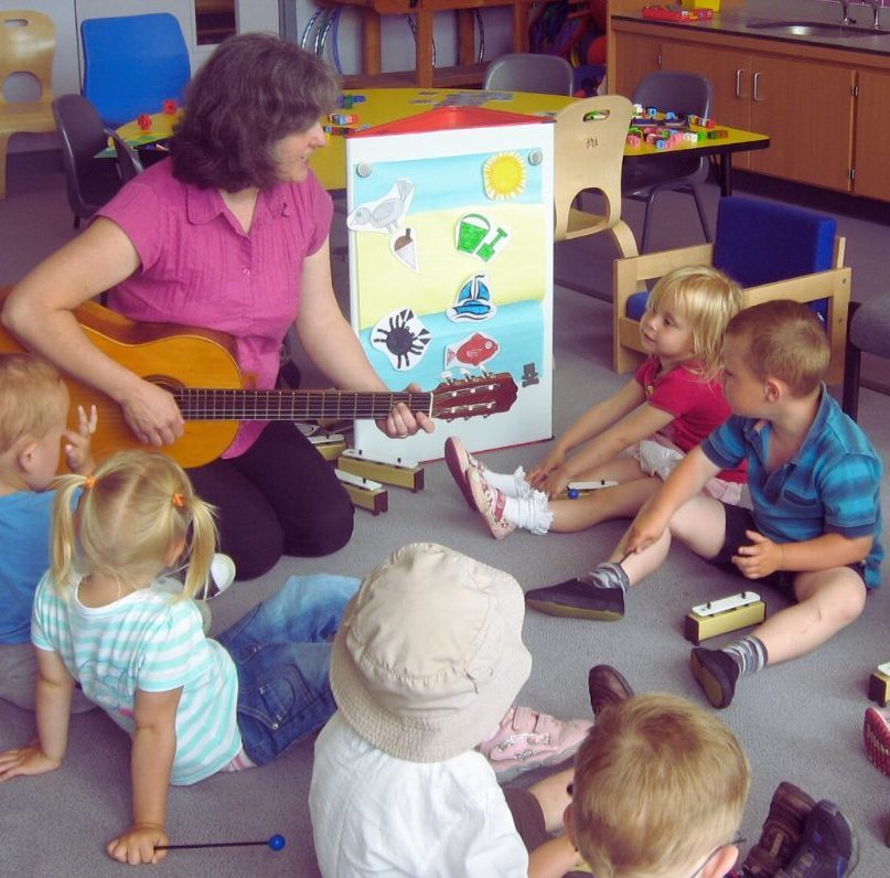 A woman plays guitar to a group of small children. They each have chime bars and are sitting on the floor. There is a board with a seaside image on it.