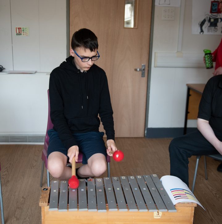 A seated boy uses red beaters to play a glockenspiel. A Figurenotes score sits on the side of the instrument.