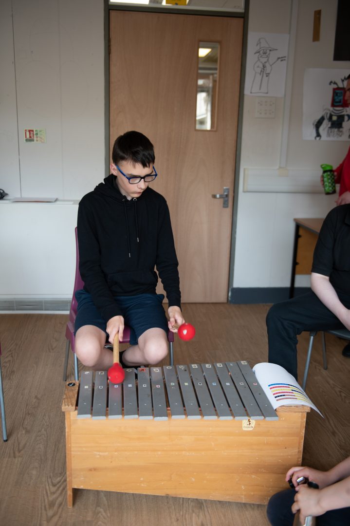 A seated boy uses red beaters to play a glockenspiel. A Figurenotes score sits on the side of the instrument.