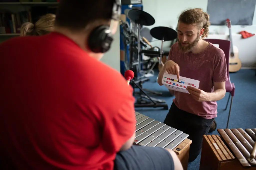 A man holds a Figurenotes score and points to it. We look over the shoulder of someone playing a glockenspiel with red beaters.