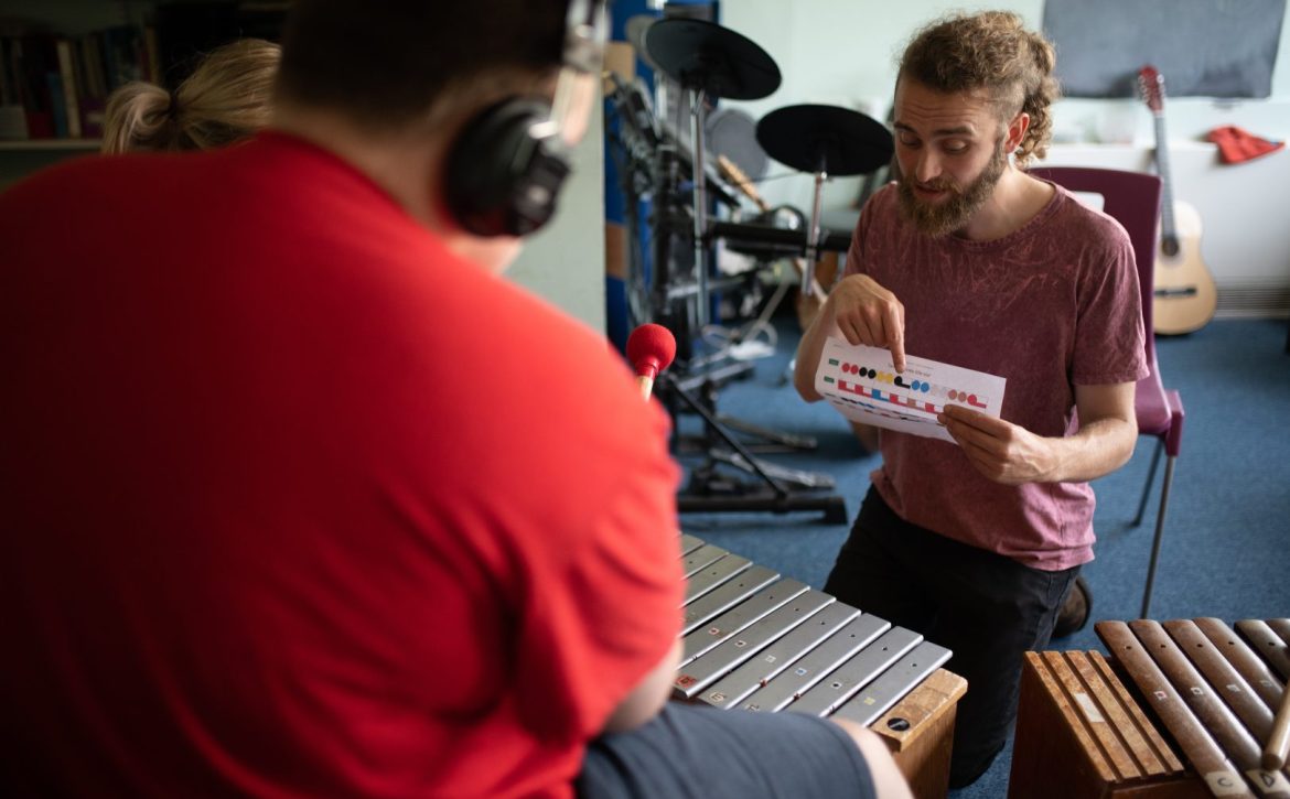 A man holds a Figurenotes score and points to it. We look over the shoulder of someone playing a glockenspiel with red beaters.