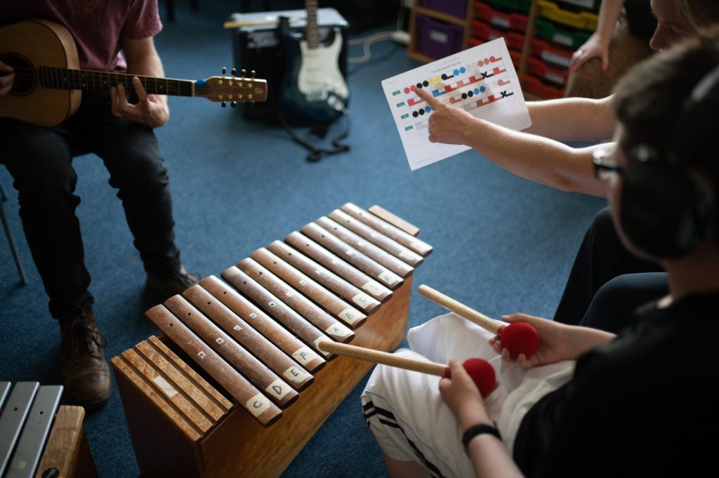 Arms hold and point to a Figurenotes score. Another set of hands play the xylophone using wooden beaters. There are Figurenotes stickers on the instrument.