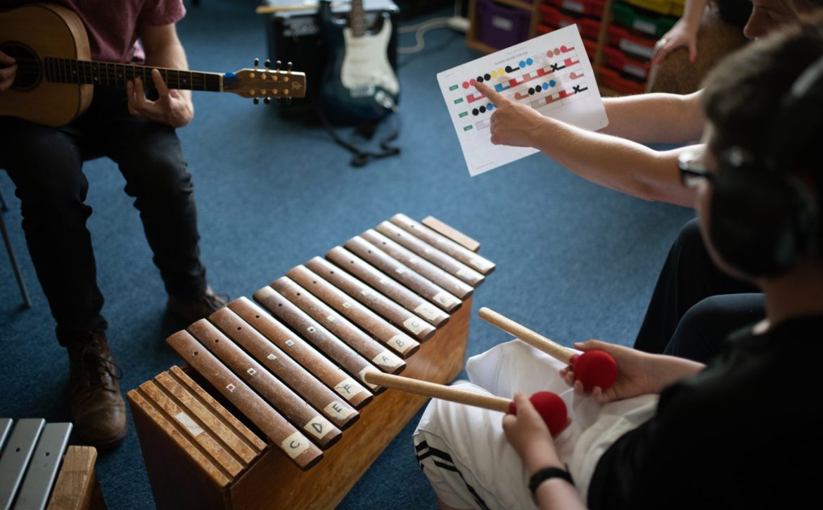 Arms hold and point to a Figurenotes score. Another set of hands play the xylophone using wooden beaters. There are Figurenotes stickers on the instrument.
