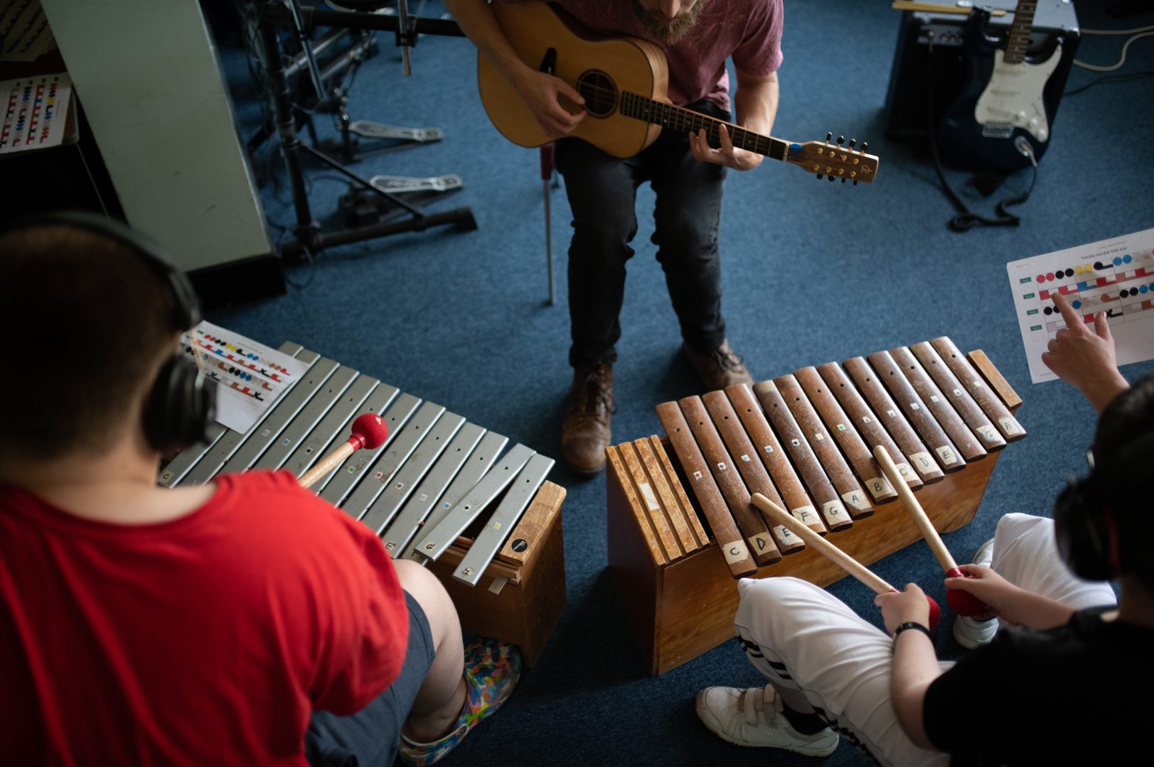 A birds-eye view of a glockenspiel played with red beaters, a xylophone played with wooden beaters, and a guitar being played.