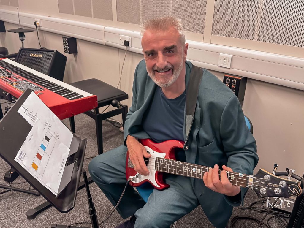 Tommy Sheppard plays a red and white shorty bass in a studio with a synth behind him. He smiles at the camera. To the side is a music stand with Figurenotes notation on it. 