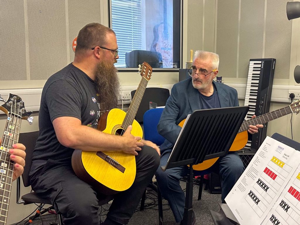 2 bearded men, Bas and Tommy, chat together. Both have guitars ready to play. Bas is explaining the Figurenotes guitar.