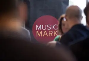 Crowd of people at a conference chatting. Slightly blurred focus. Music Mark logo in the centre. Logo is a maroon circle with the words Music Mark written in white and yellow.