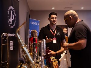 Scene from a stall at a music conference. Brass instruments are visible and two men in black t shirts are enthralled in conversation. 
