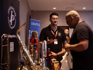 Scene from a stall at a music conference. Brass instruments are visible and two men in black t shirts are enthralled in conversation.