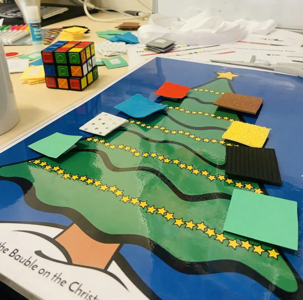 A laminated image of a Christmas tree sits on a desk with different textured squares attached in Figurenotes colours.
