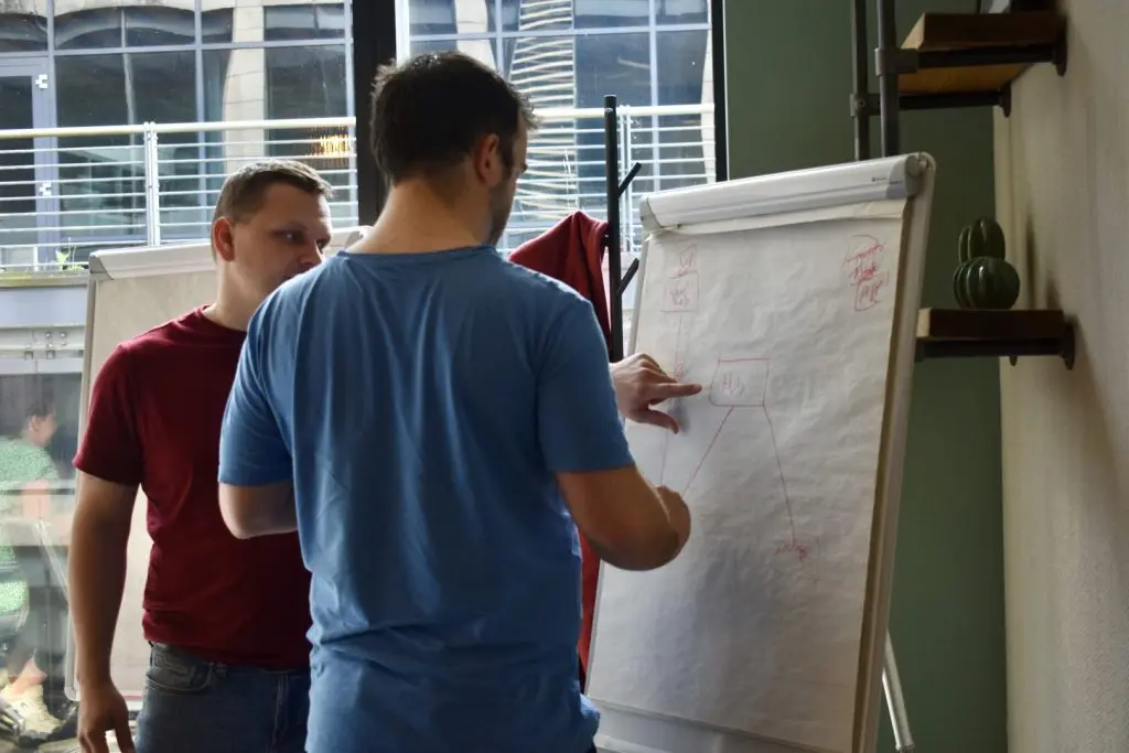 2 men in discussion about what they are writing on a board. One man points to the diagram while the other looks on.