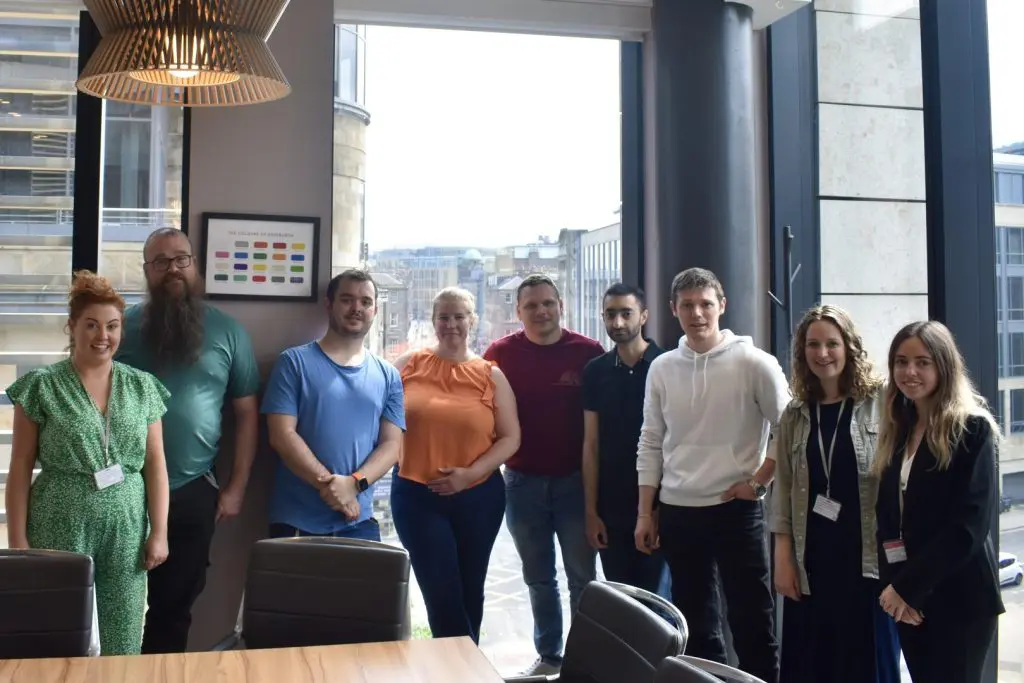 Group of nine people smiling, standing side by side in a professional manner, in front on a large windowed wall.