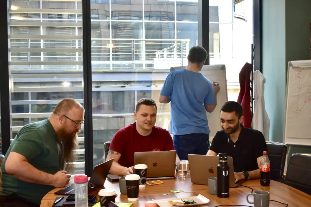 3 men sit at a desk working on computers. 1 man in a blue t-shirt writes on a board in the background.