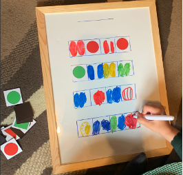 A whiteboard with 4 Figurenotes bar on it. A child's hand holds a pen and is drawing Figurenotes rhythms into the bars using different colours.
