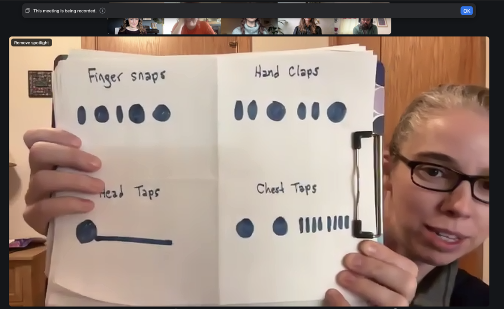 A woman with glasses holds up a piece of paper to the camera on Zoom. On it are hand drawn Figurenotes rhythms with body percussion instructions - Finger snaps, hand claps, head taps, chest taps. 5 people watch at the top of the screen.