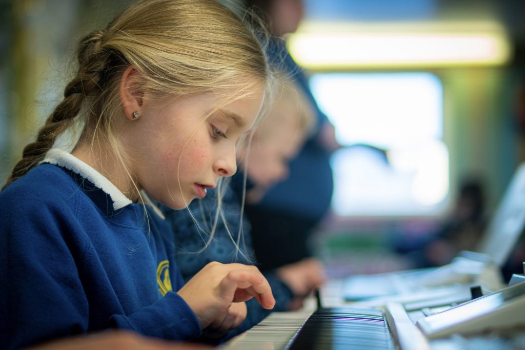 blonde child with pigtails and a blue jumper on, playing keyboard.