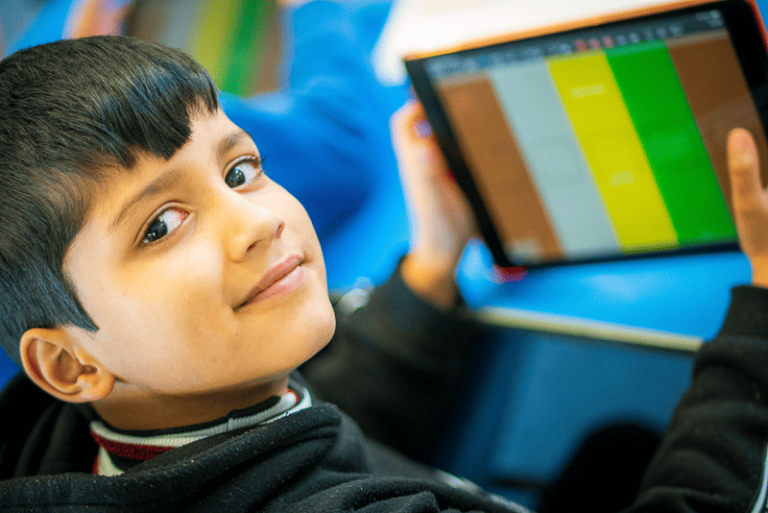 A boy gently smiles at the camera. He holds an ipad with coloured bars on the screen in Thumbjam