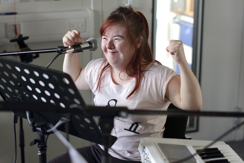 A girl is surrounded by musical equipment. She holds her fists aloft in a triumphant movement.