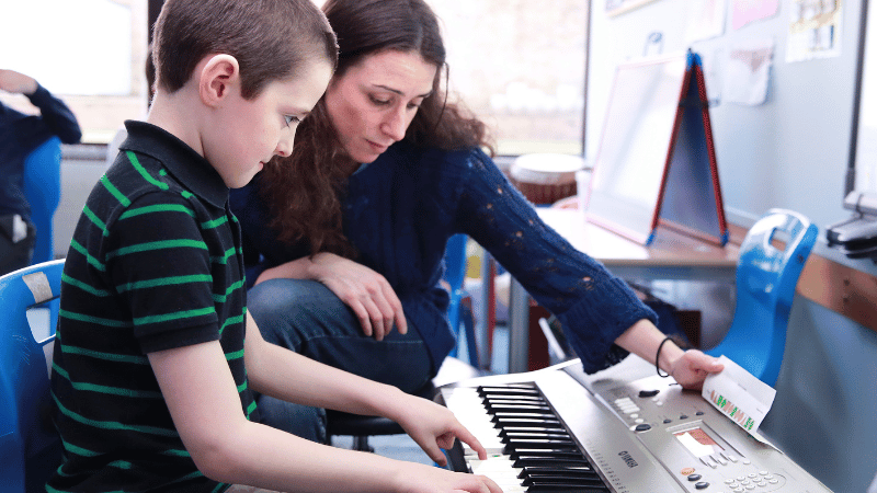 A boy plays a keyboard. He is concentrating hard, while his teacher sits calmly looking at the keys.