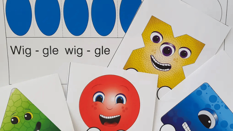 Cute monsters made from the Figurenotes shapes of square, triangle, circle, and cross. A Figurenotes score sits behind them with the words 'Wiggle Wiggle' visible