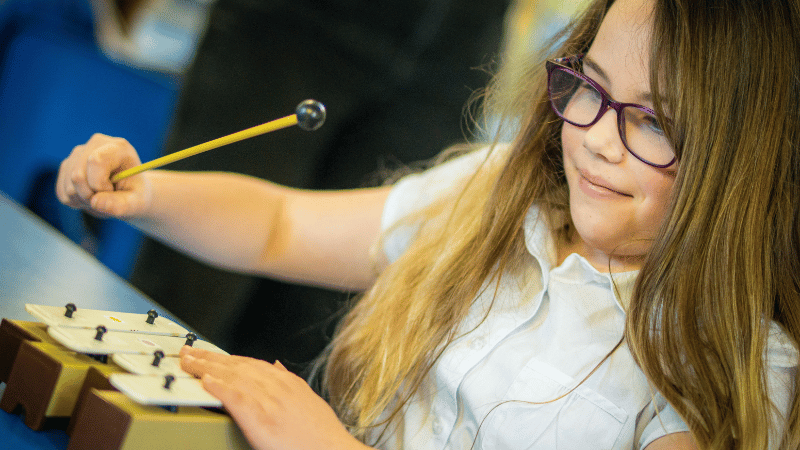 A girl with long hair and glasses is playing the chime bars.