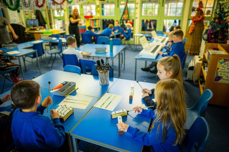 A primary school classroom. The children sit at blue tables with chime bars and beaters.