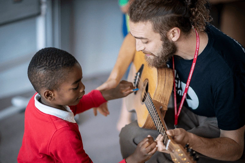 A man holds a guitar and shapes the chords, while a young boy strums the strings.