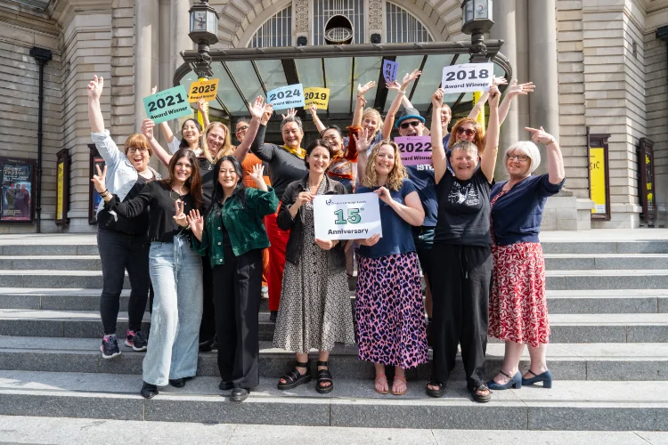 A group of people cheer. They are holding signs with different years on them, such as '2022 Award Winner'. 2 women in the middle hold a sign saying '15th Anniversary'